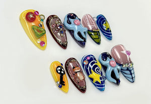 A collection of ten almond-shaped, embellished artificial nails. Designs include a key, an octopus, and a star, with colors like yellow, blue, and brown. Each nail is adorned with gems and intricate details.