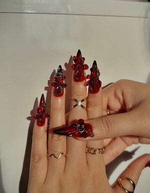 Red acrylic nails with intricate floral and vine-like embellishments. The nails are long and pointed, with a glossy finish. Gold rings adorn the fingers, adding to the overall aesthetic. The design is bold and eye-catching.