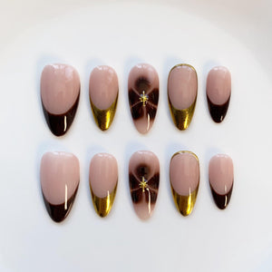 A set of ten artificial nails in various shapes. The nails feature a light pink base with dark brown tips, gold accents, and a dark brown swirl design with a gold star. The nails are arranged on a white surface.