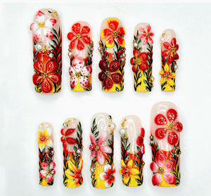 A set of ten artificial nails with a floral design. Each nail features a base of yellow, orange, and red, with 3D flowers in red, white, and pink. Some nails have pearl and rhinestone accents.