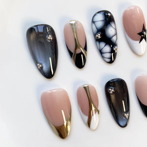 A collection of artificial nails in various designs. Some nails are black, gray, or nude with metallic accents. Others feature a black and white marbled pattern or a star design. Each nail has a glossy finish and is oval-shaped.