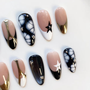 A collection of oval-shaped artificial nails. Designs include black and white patterns, gold metallic accents, and a star design. Some nails feature small, silver-colored embellishments.