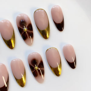 A set of almond-shaped false nails. The nails feature a nude base with gold metallic tips and accents. Some nails have a dark brown swirl design with a gold star-shaped embellishment. The nails are arranged on a white surface.