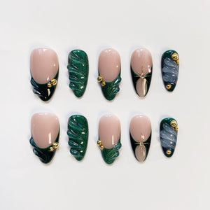 A set of ten artificial nails in various designs. The nails feature a nude base with dark green and gold accents, along with a few nails with a silver star design and a swirling blue-gray pattern. The nails are oval-shaped.