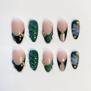 A set of ten artificial nails in various designs. The nails feature a nude base with dark green and gold accents, including a textured wave design and starburst embellishments. Some nails have a dark green French tip.