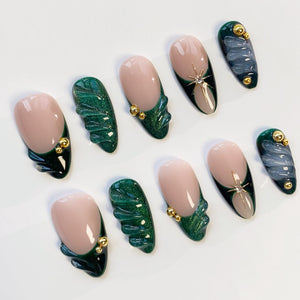 A set of ten artificial nails with various designs. Some nails feature a nude base with a dark green French tip and gold accents. Others have a swirling blue and gray design, and some have a green textured design.
