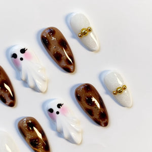 A collection of artificial nails with various designs. Some nails are white with gold accents, others are brown with a leopard print, and some feature a cartoon ghost design with pink cheeks.