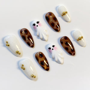 A collection of almond-shaped, decorated artificial nails. Designs include white nails with gold accents, leopard print, and small white ghost figures with pink cheeks and black eyes.