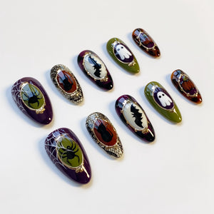A collection of Halloween-themed press-on nails. The nails feature designs such as spiders, bats, ghosts, and pumpkins. Colors include purple, green, orange, and black, with gold accents. The nails are oval-shaped and have a glossy finish.