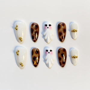 A set of ten press-on nails. The nails feature a variety of designs, including white with gold accents, brown leopard print, and white ghost designs with pink cheeks. The nails are oval-shaped.