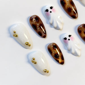 A collection of artificial nails with various designs. Some nails are white with gold embellishments, others feature a brown and gold pattern, and some are shaped like cartoon ghosts with pink cheeks.
