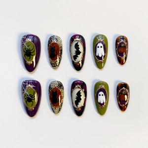 A set of ten Halloween-themed press-on nails. Each nail features a different design, including a spider and web, a black cat, a bat, a ghost, and a pumpkin. The nails are oval-shaped and painted in various colors, including purple, green, and orange.