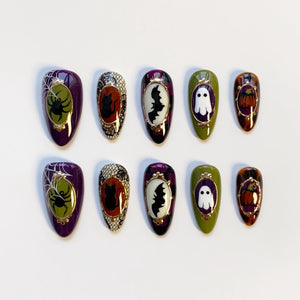 A set of ten Halloween-themed press-on nails. Each nail features a different design, including a spider and web, a black cat, a bat, a ghost, and a pumpkin. The nails are oval-shaped and painted in various colors, including purple, green, and orange.