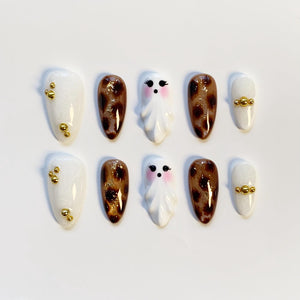 A set of ten press-on nails in various designs. The nails feature white, brown leopard print, and ghost designs. Some nails have gold bead accents. The nails are oval-shaped and are arranged on a white surface.