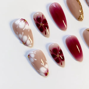 A collection of almond-shaped artificial nails. Some nails feature a gradient of red to pink, while others showcase a floral design with white petals, red accents, and gold embellishments. The nails are set against a white background.