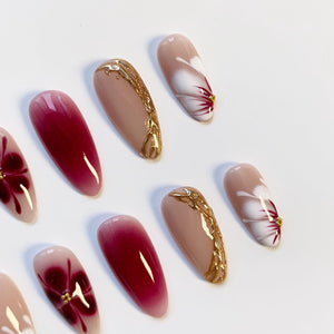 A collection of artificial nails in various designs. Some nails are a gradient of red to clear, others feature a white flower design with red accents, and some have a nude base with gold embellishments. Each nail is oval-shaped.
