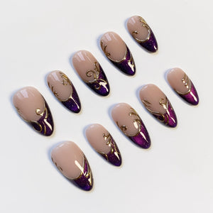 Set of ten almond-shaped false nails. Each nail features a nude base, a deep purple glitter tip, and gold filigree accents. The nails are arranged on a white surface, showcasing their elegant design.