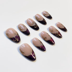 Set of artificial nails with a nude base, a dark purple glitter gradient, and gold filigree accents. The nails are oval-shaped and arranged on a white surface. The design is elegant and suitable for special occasions.