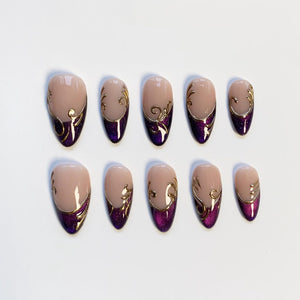 Set of ten almond-shaped false nails. Each nail features a nude base, a deep purple glitter accent at the base, and gold filigree detailing. The nails are arranged on a white surface.