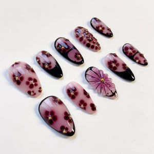 A collection of artificial nails in various designs. The nails feature a glossy finish with shades of pink, purple, and black. Some nails are adorned with small gold embellishments and floral patterns, while others have a flower-shaped design.