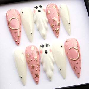 A set of ten long, almond-shaped artificial nails. The nails feature a variety of designs, including a pink base with gold stars and moons, white nails with silver studs, and white ghost-shaped nail art.