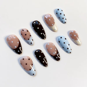 A collection of ten artificial nails in various designs. Some nails are light blue with black polka dots, others are brown with white polka dots, and some have a beige and white design with a dark brown tip.
