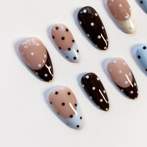 A collection of oval-shaped false nails. The nails feature a variety of designs, including solid dark brown, light pink with dark brown polka dots, and light pink with a light blue tip and dark brown polka dots. The nails are arranged on a white surf