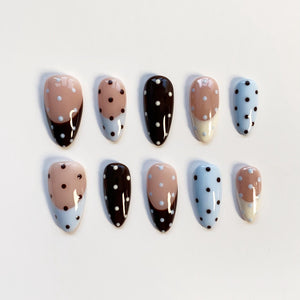 A set of ten almond-shaped false nails. The nails feature a variety of designs, including solid colors, polka dots, and color-blocked patterns. Colors include light blue, brown, and beige.