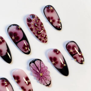A collection of artificial nails with a floral design. The nails feature a gradient of pink to dark purple, with gold accents. Some nails have a 3D flower design, while others have gold studs.