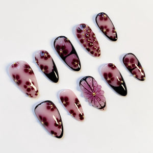 A collection of artificial nails with various designs. The nails feature a gradient of pink and purple hues, adorned with floral patterns and small gold embellishments. Some nails have a black base, while others showcase a 3D flower design.