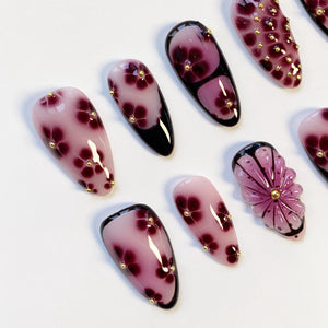 A collection of artificial nails with various designs. Some nails feature a gradient of pink and purple with dark red floral patterns and gold accents. Others have a black base with a floral design.