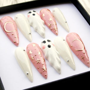 A set of ten long, pointed false nails. The nails feature a variety of designs, including white, pale pink with gold stars, and pink with a gold crescent moon. Some nails have small ghost figures.
