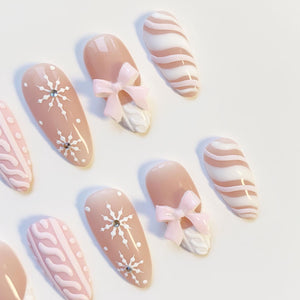 A set of almond-shaped false nails in various designs. Some nails feature white snowflake patterns with rhinestones, others have pink bows and white textured accents, and some have pink and white wavy stripes.