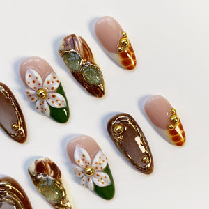 A collection of artificial nails with various designs. Some nails feature a nude base with gold accents, while others showcase intricate floral patterns with white petals and gold centers. Other nails have a brown marble effect.