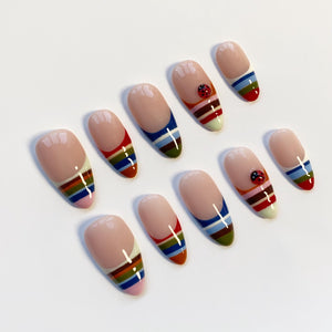 A set of ten artificial nails with a nude base and colorful striped designs. Some nails feature a small ladybug accent. The stripes include red, blue, green, and yellow, creating a vibrant and playful look.