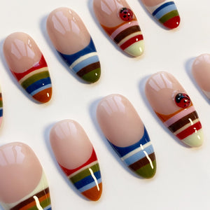 Oval-shaped false nails with a light pink base and colorful striped designs. Some nails feature a small ladybug accent. The stripes include red, orange, green, blue, and brown. The nails have a glossy finish.