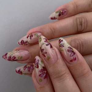 Almond-shaped nails with a floral design. The nails feature a nude base with hand-painted burgundy flowers, green leaves, and gold accents. The design is delicate and elegant.
