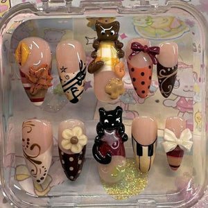 A set of ten artificial nails with various designs. The nails feature a range of colors, including nude, brown, orange, and white, and are adorned with decorative elements such as bows, flowers, and animal figurines. The nails are displayed in a clea