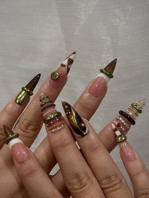 A set of long, pointed artificial nails. The nails feature a variety of designs, including brown and white checkered patterns, gold accents, and decorative rings with pearls and gems. The nails have a natural pink base with white tips.