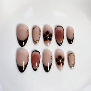 Set of ten almond-shaped false nails. The nails feature a nude base with various designs, including dark brown tips, floral accents, and gold embellishments. Some nails have a dark brown gradient, while others have gold swirls.