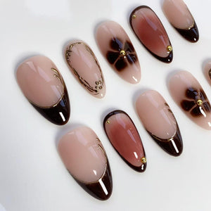 A collection of almond-shaped artificial nails. The nails feature a variety of designs, including a dark brown French tip with gold trim, floral patterns, and gold embellishments. The base color is a nude pink.