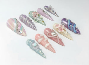 An assortment of long, pointed artificial nails in various pastel colors. Some nails feature decorative elements like pearls, rhinestones, and floral designs. The nails are arranged on a white surface.