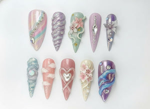A collection of ten long, almond-shaped artificial nails. Each nail features unique designs, including swirls, gems, flowers, and cloud-like accents. Colors range from pastel pinks and blues to purples and greens, with iridescent and metallic finishe