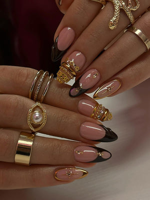 Close-up of manicured nails with a pink base and black tips. The nails are adorned with gold rings, including a snake ring and rings with pearls and gems. The nails have a French tip design.