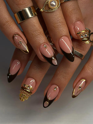 A close-up of manicured nails with a light pink base, dark brown tips, and gold accents. Some nails feature gold starburst designs, while others have gold embellishments and rings. The nails are long and almond-shaped.