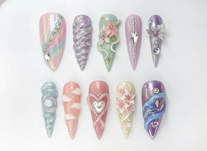 An assortment of ten long, almond-shaped artificial nails. Each nail features unique designs, including iridescent finishes, sculpted elements, and embellishments like rhinestones, pearls, and floral accents. Colors range from pastel pinks and purple