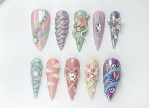 An assortment of ten long, almond-shaped artificial nails. Each nail features a unique design, including iridescent colors, 3D floral accents, and sparkling rhinestones. The nails are arranged on a white surface.