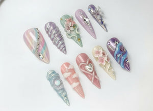 A collection of ten long, almond-shaped artificial nails. Each nail features unique designs, including pastel colors, pearls, floral accents, and 3D embellishments. The nails are arranged on a white surface.