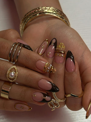 A hand adorned with gold rings and a gold bracelet. The nails are painted with a French manicure design, featuring a nude base with black tips and gold accents. Some nails have gold embellishments.