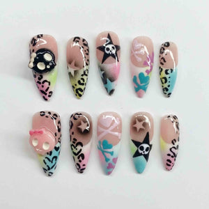 A set of ten long, pointed artificial nails. Designs include leopard print, skulls, stars, hearts, and crossbones in black, pink, blue, and yellow. Some nails feature 3D skull embellishments.